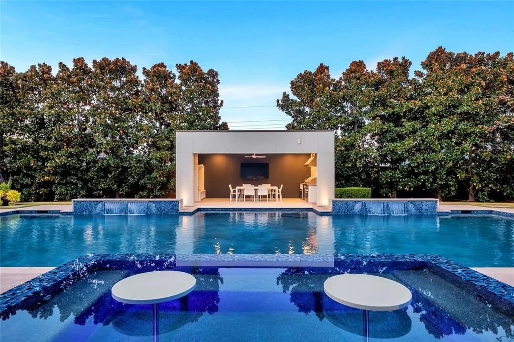 Modern backyard with a large swimming pool, a covered outdoor dining area, and lush green trees in the background.