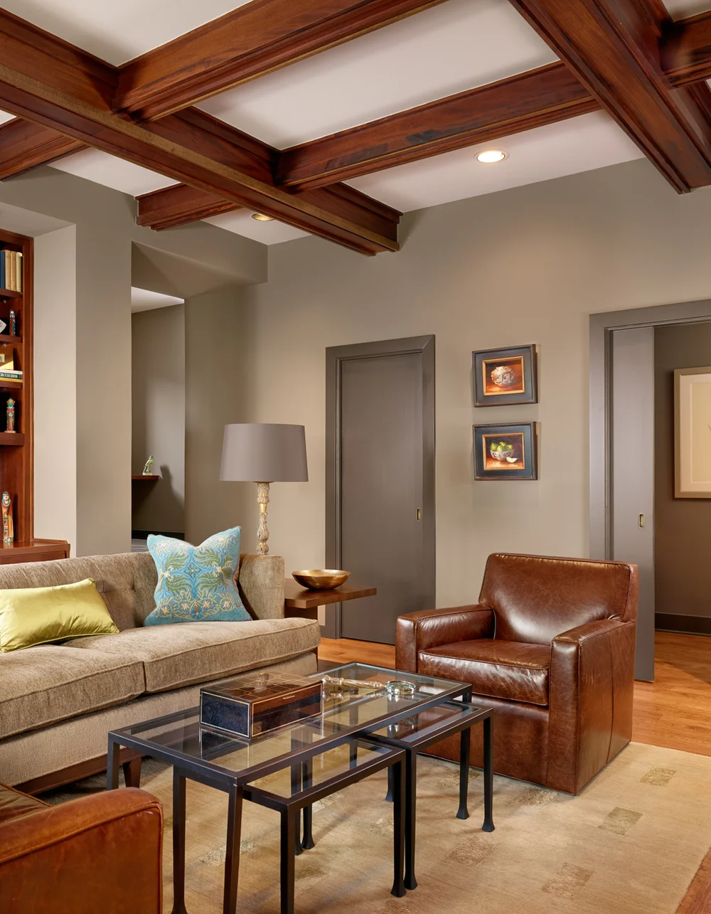 Living room with beige couch, brown leather armchair, glass coffee tables, wooden ceiling beams, walls with framed pictures, table lamp, and a beige area rug.