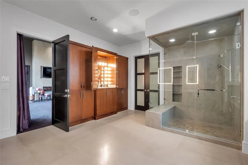 Modern bathroom with a glass-enclosed shower area, built-in shelving, and a wooden cabinet with a lit vanity mirror and personal items on top.