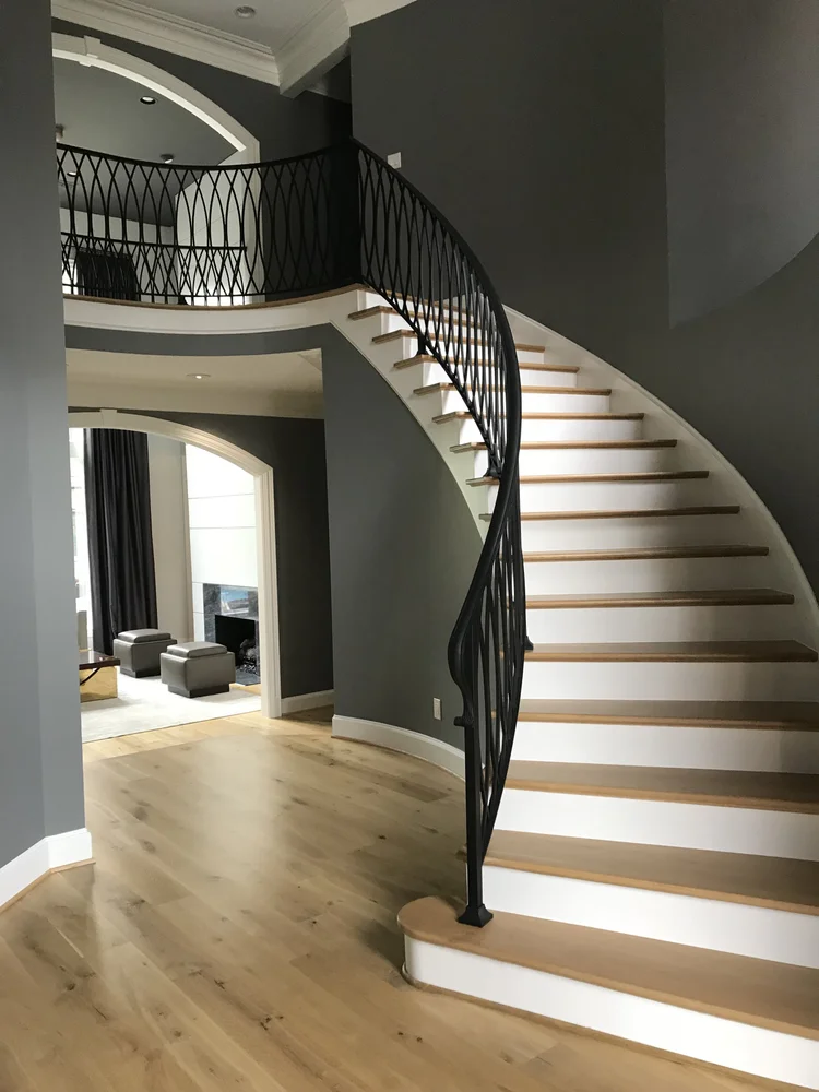 Interior view of a modern home featuring a curved staircase with black railing and wooden steps, leading to an upper level. The space has gray walls and light wooden flooring, with a living area visible in the background through a large arched doorwa
