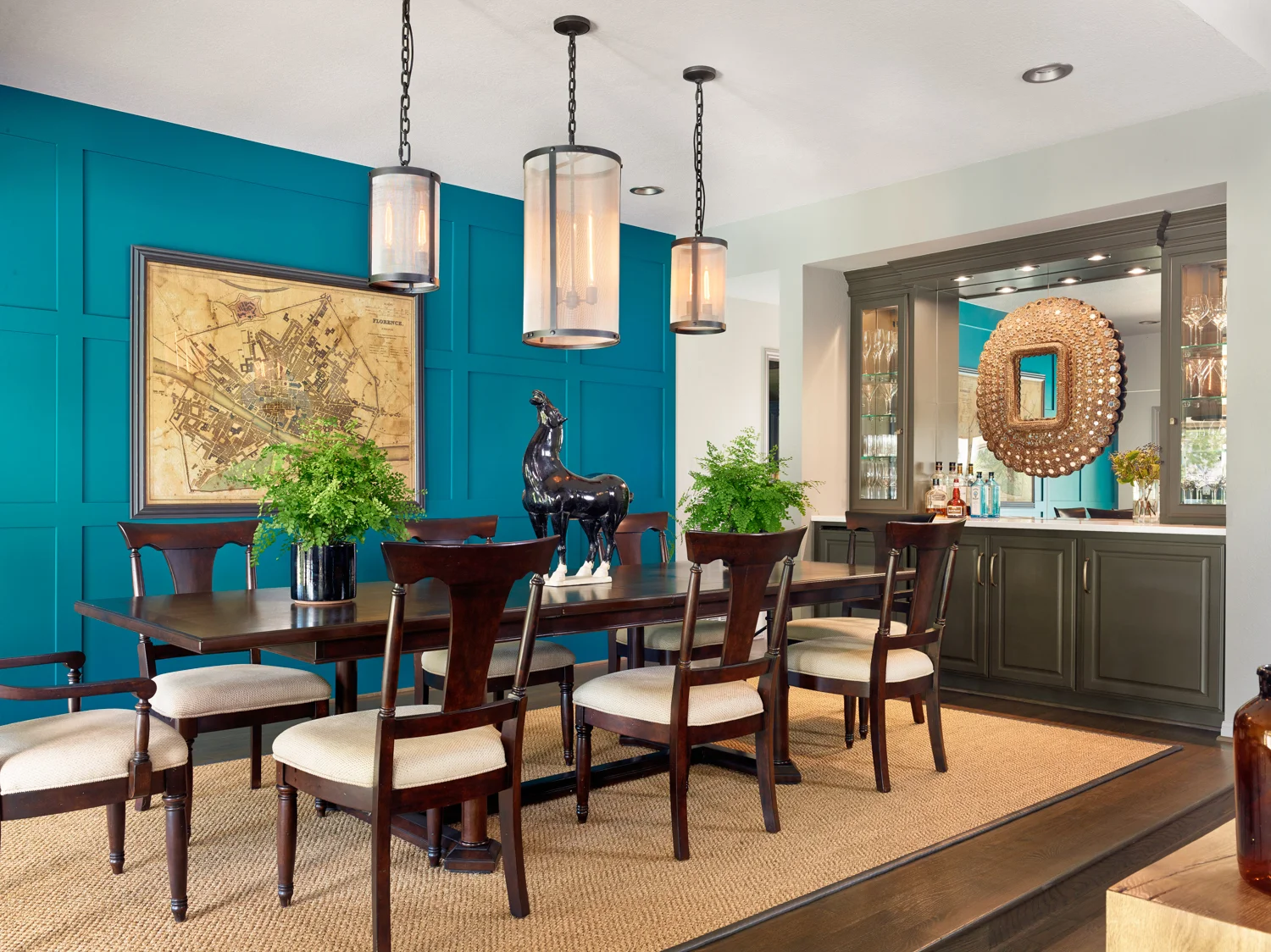 Dining room with a dark wood table, beige upholstered chairs, teal accent wall with a map, hanging pendant lights, green plants, and a bar area with glassware and bottles, decorated with a large mirror.