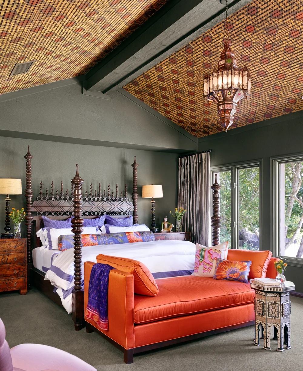 A bedroom with a four-poster bed, nightstands, an orange velvet bench at the foot of the bed, and a decorative ceiling with a large oriental lamp hanging from the ceiling. The room has large windows with curtains and various decorative items such as 
