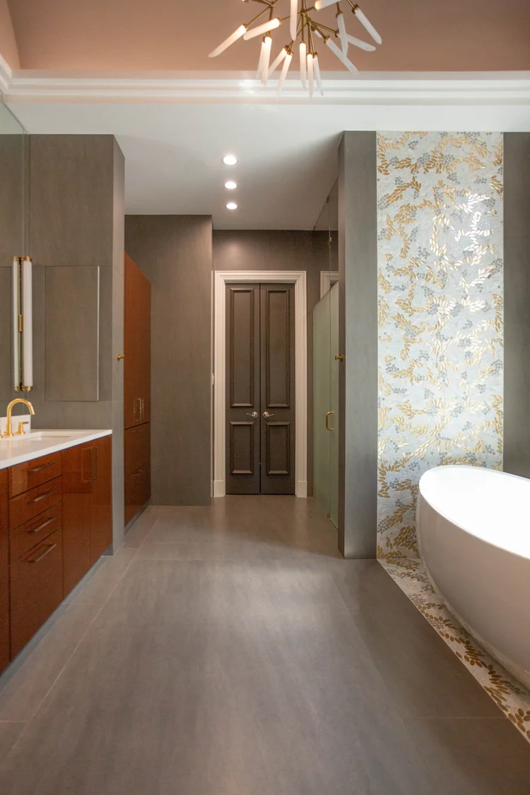 Modern bathroom with gray and wood cabinetry, gold fixtures, a free-standing bathtub, patterned wallpaper, and ceiling lighting