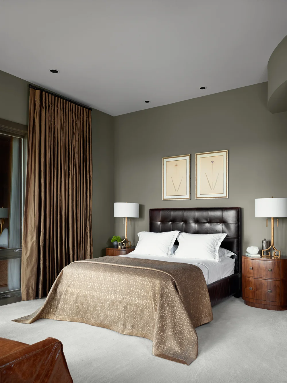 A bedroom with a dark leather headboard, two white pillows, a beige patterned bedspread, and two nightstands with lamps. The walls are painted light gray, and there are two framed pictures hanging above the bed. A large brown curtain covers a window 
