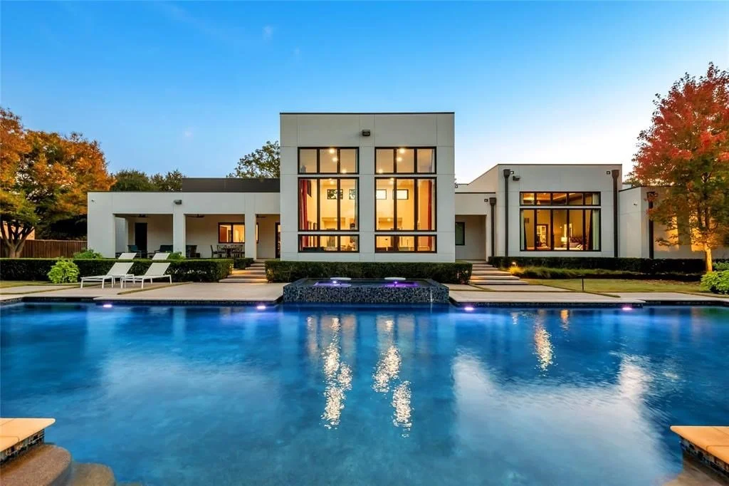 Modern house with large glass windows, outdoor pool, green lawn, and trees with fall foliage at dusk.