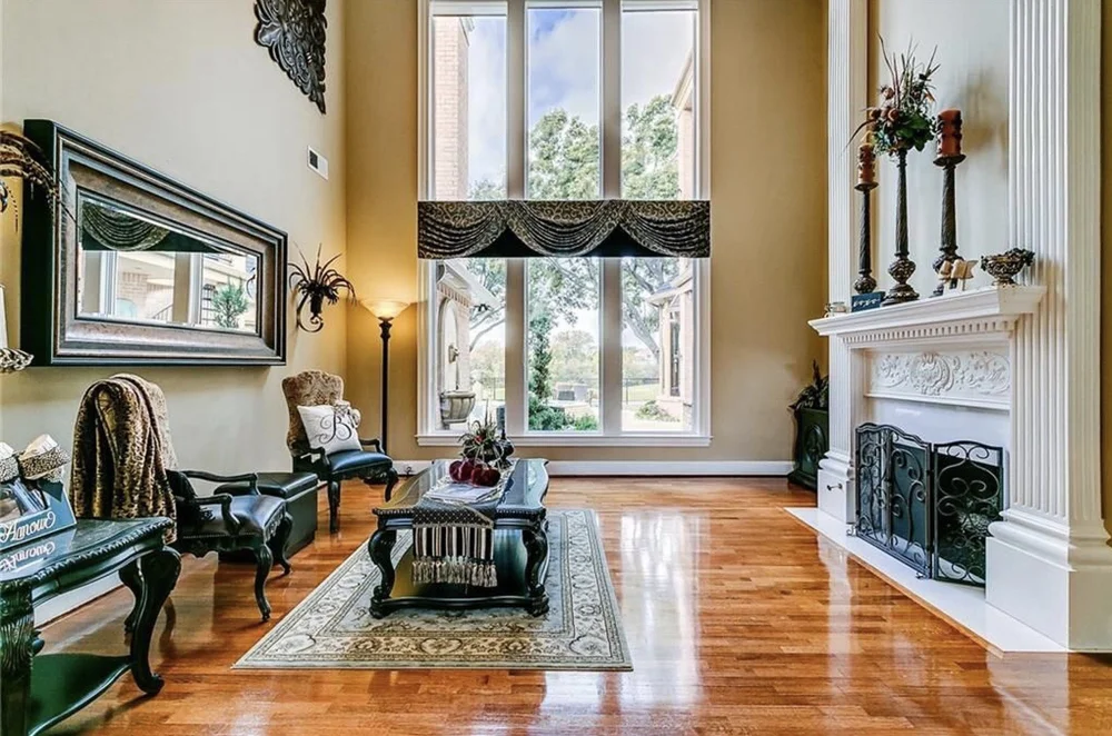 Bright living room with large window, black leather chairs, a patterned area rug, ornate white fireplace with decorative candles, and a mirror on the wall.