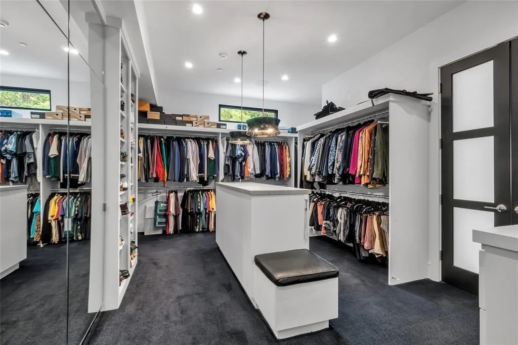 A walk-in closet with white shelves filled with hanging clothes, a central white island, black cushioned bench, black door, and black carpeted floor. Small windows allow natural light.