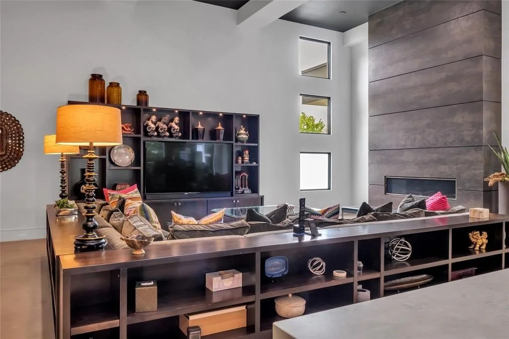 Modern living room with a black entertainment center, a large flat-screen TV, a variety of decorative items, a gray sofa with multi-colored pillows, a fireplace on the gray wall, and tall narrow windows.