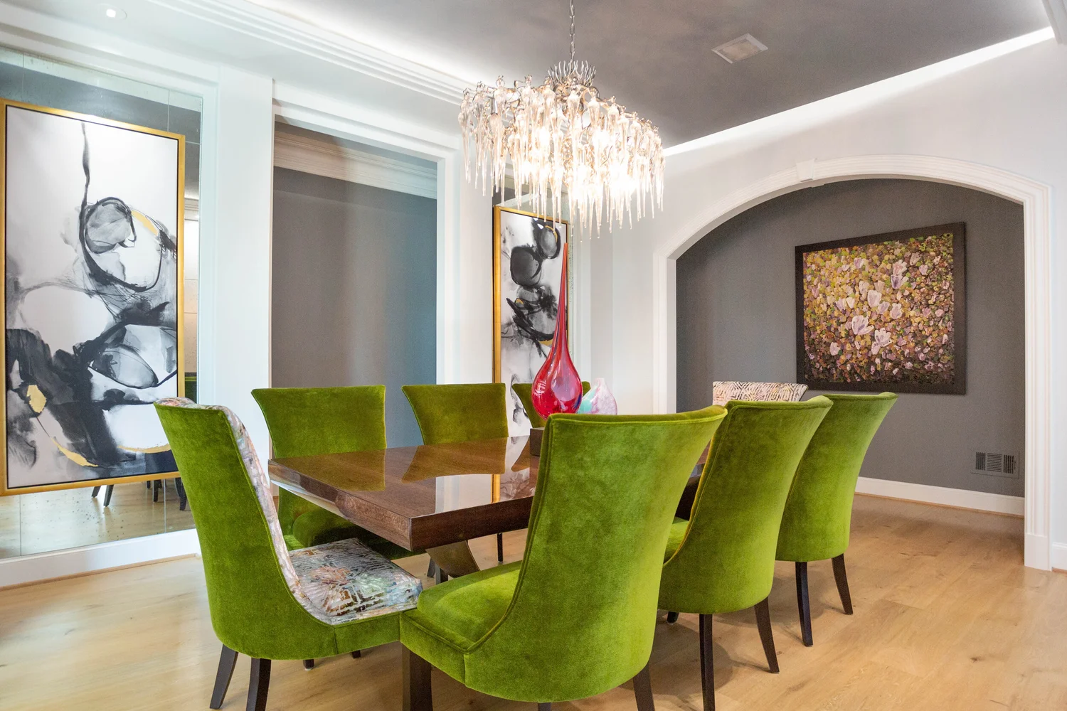 Dining room with a wooden table surrounded by six green velvet chairs, a chandelier hanging above, colorful vases on the table, and abstract paintings on the walls.
