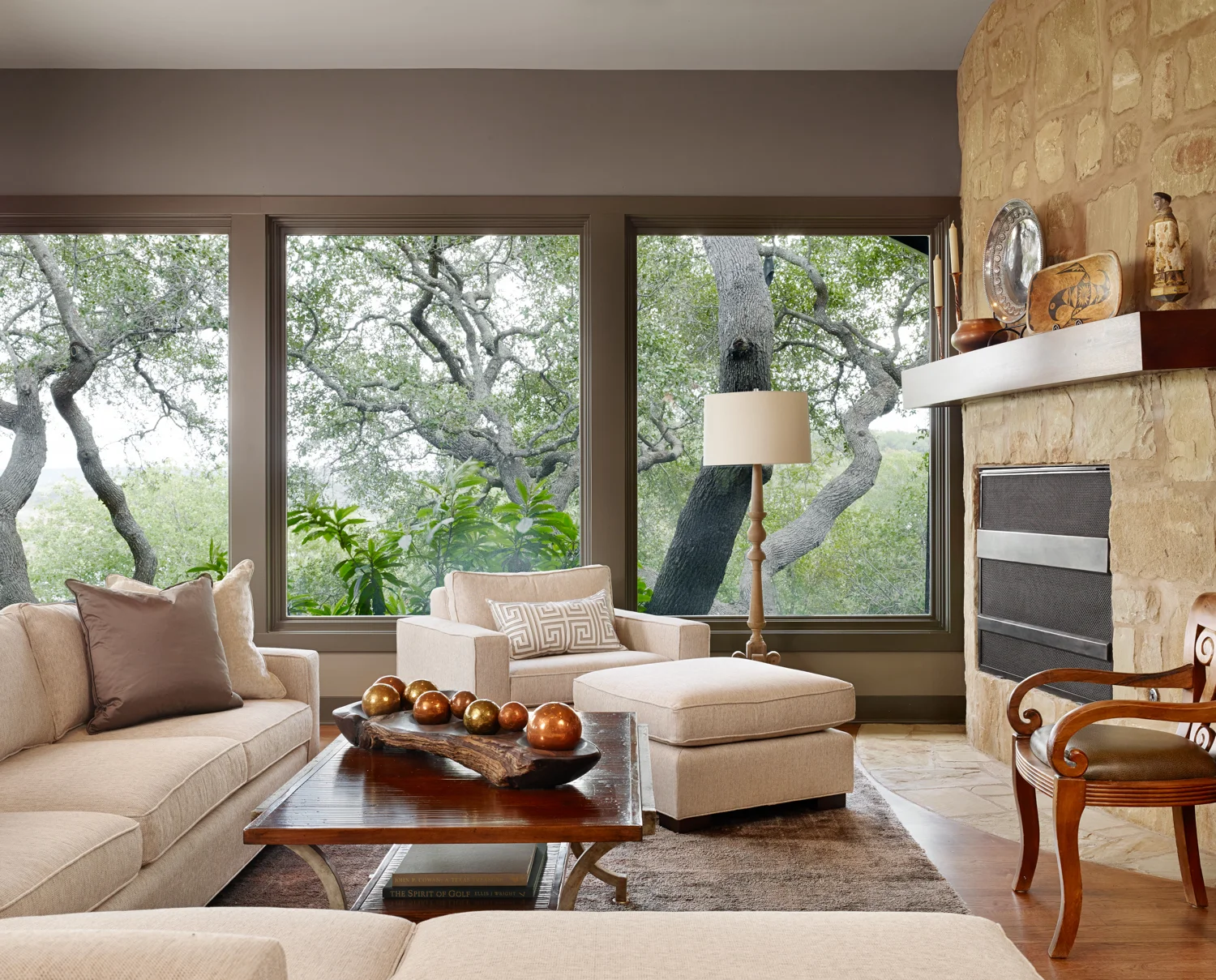 Living room with large windows showing trees outside, beige sofas, a wooden coffee table with decorative gold and copper spheres, a floor lamp, fireplace with stone surround, and various decorative items on the mantel.
