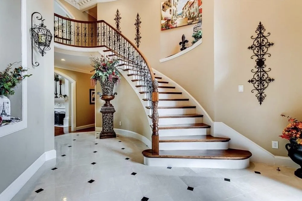 Interior of a house with a curved staircase featuring wooden steps and black ornate iron railing. Decor includes floral arrangements on a tall pedestal, wall-mounted wrought iron art, and a large painting of a street scene. The flooring is white with