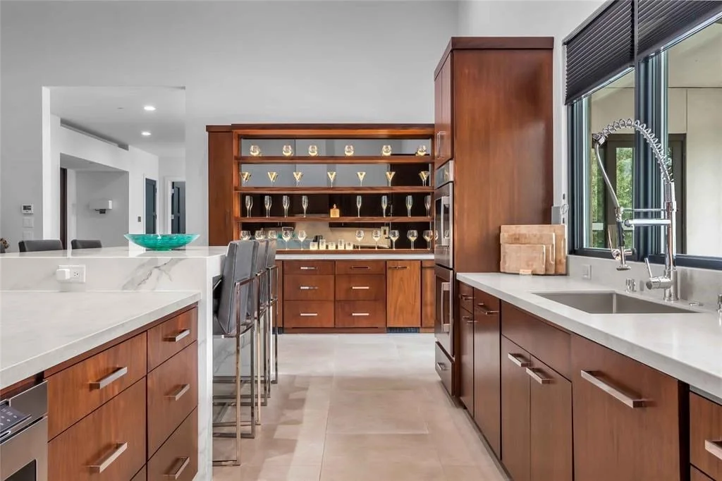 Modern kitchen with white marble countertops, wooden cabinets, a large window with a black blind, and a built-in wooden shelf displaying glassware and decorative items.