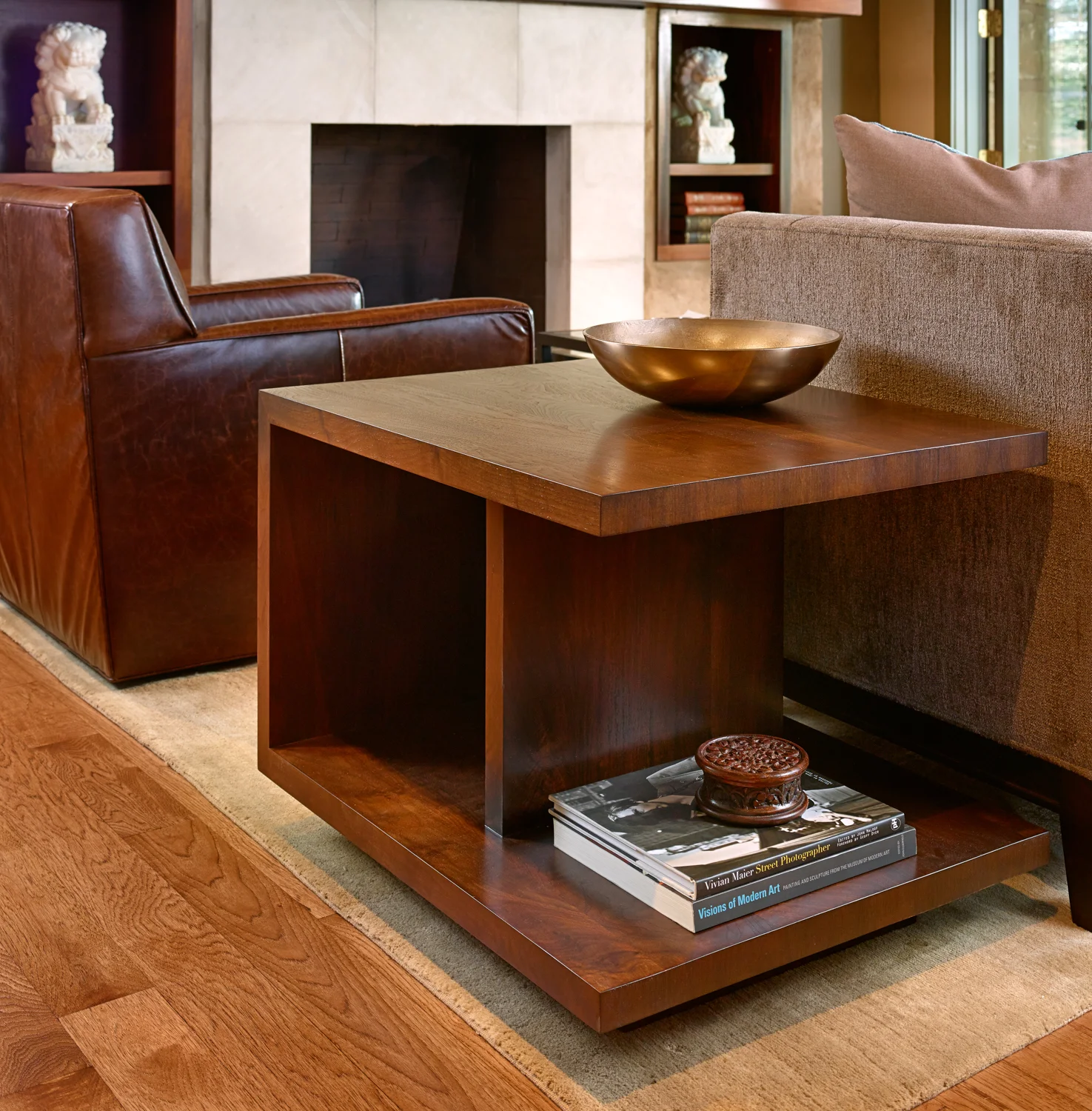 A living room with a wooden end table holding a gold decorative bowl and a stack of art books. The background features a tan sofa, a stone fireplace, and built-in shelves with decorative sculptures and books.