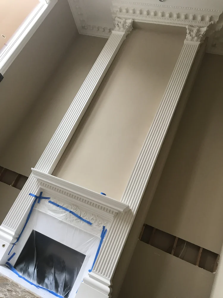 A fireplace with ornate white moulding surrounded by two fluted white columns, with decorative crown moulding at the top. The fireplace opening is covered with plastic and taped off in preparation for painting or renovation.