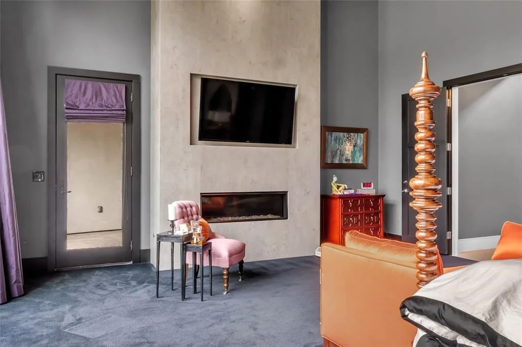 Living room with gray walls, purple curtains, a pink armchair with a small side table, a large flat-screen TV mounted above a fireplace, a red cabinet, and a wooden four-poster bed.
