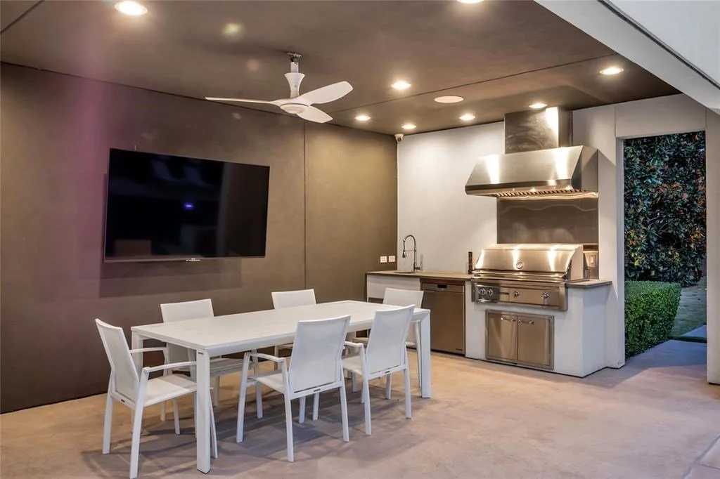 Modern outdoor dining area with a white table, white chairs, a wall-mounted TV, a stainless steel grill, and a ceiling fan.