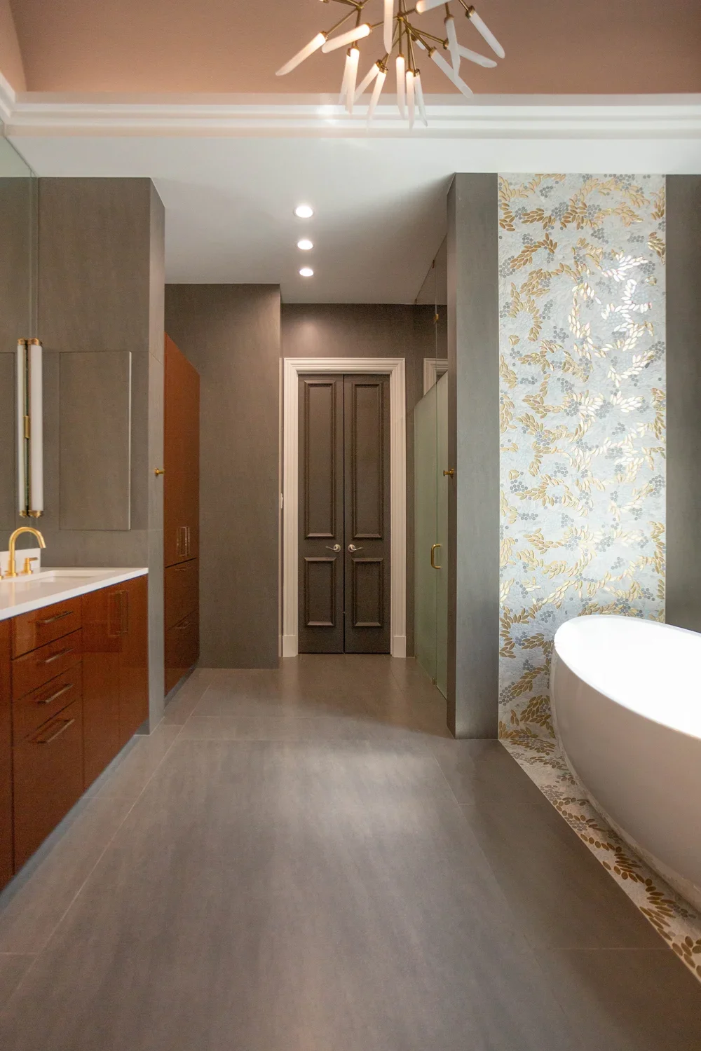 Modern bathroom with gray tiles, a white bathtub with a gold and white fixture, a vanity with a white countertop and dark wood cabinets, a decorative wall with gold and gray floral wallpaper, and a closed double door in the background.