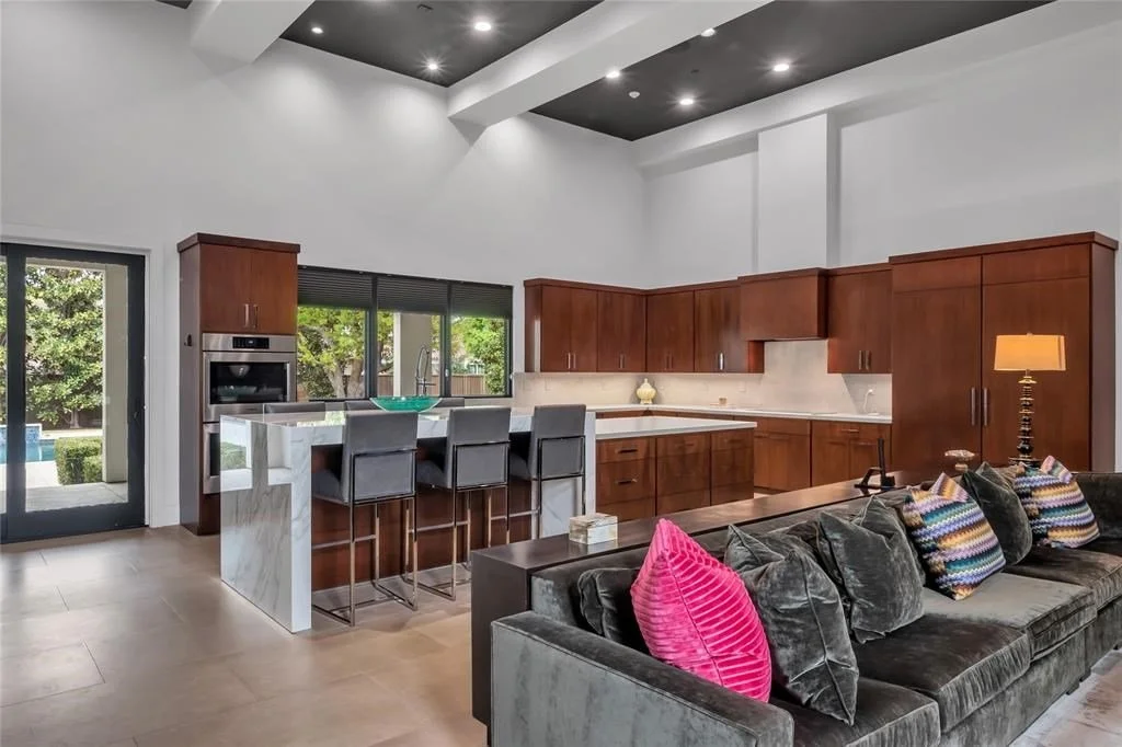 Open-concept kitchen and living room with wooden cabinets, a marble island, gray bar stools, and a gray sectional sofa with colorful pillows.