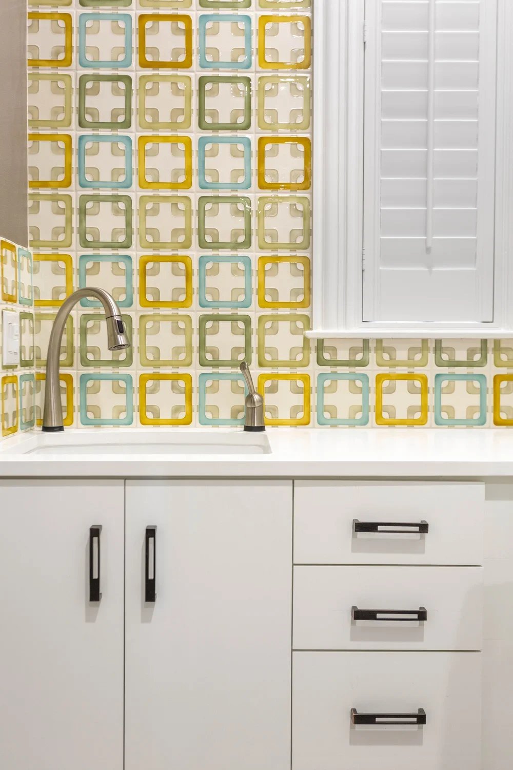 Kitchen sink with a modern faucet, white cabinets, and colorful retro geometric patterned tile backsplash in shades of yellow, green, and blue, with a window with white shutters above.