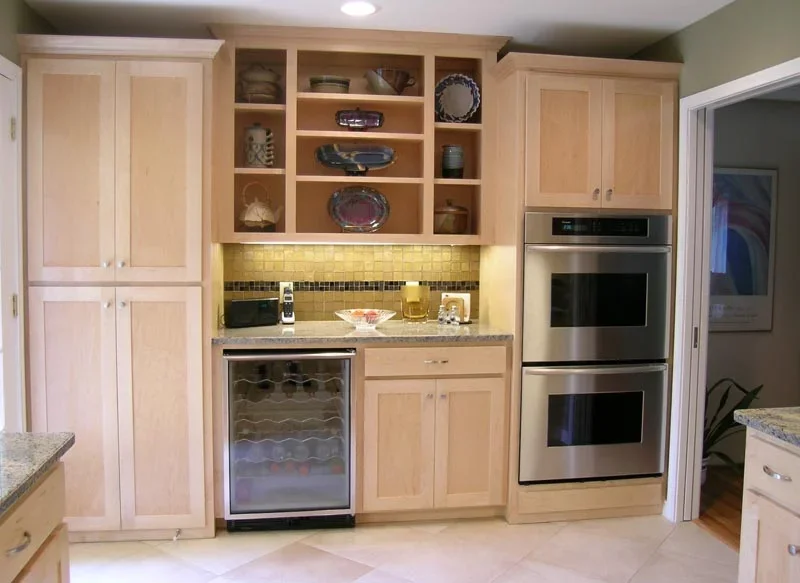 Kitchen with light wood cabinets, open shelving with decorative plates and bowls, granite countertops, a built-in wine cooler, a double oven, and a small countertop appliance.