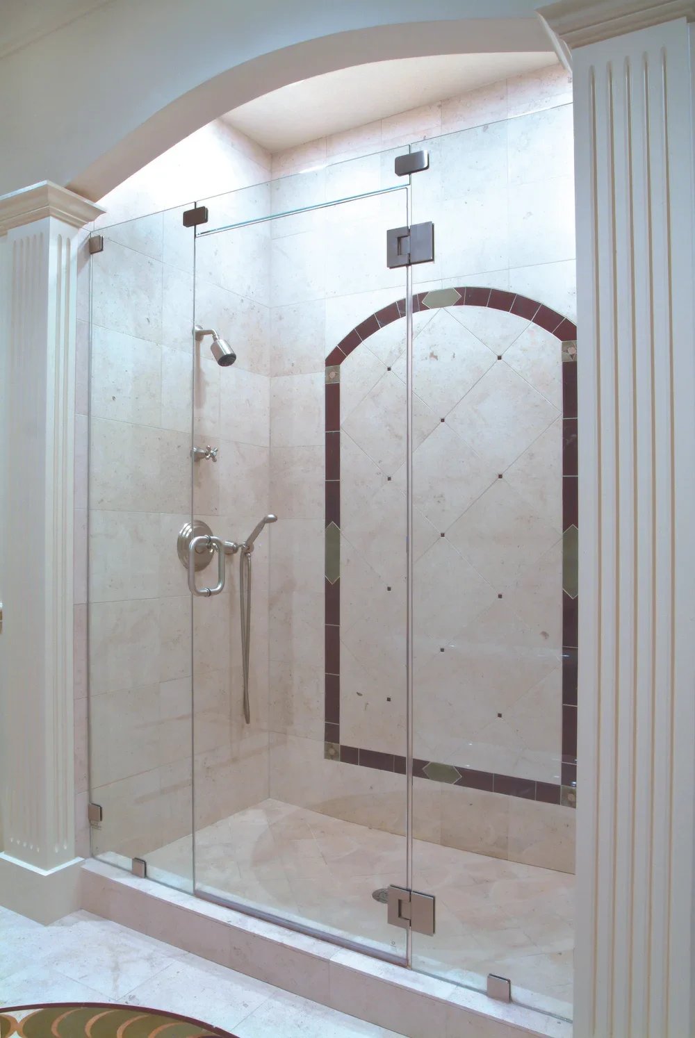 A clear glass shower with a handheld showerhead, decorative tile design on the back wall, and a partial view of a room with a patterned carpet outside the shower.