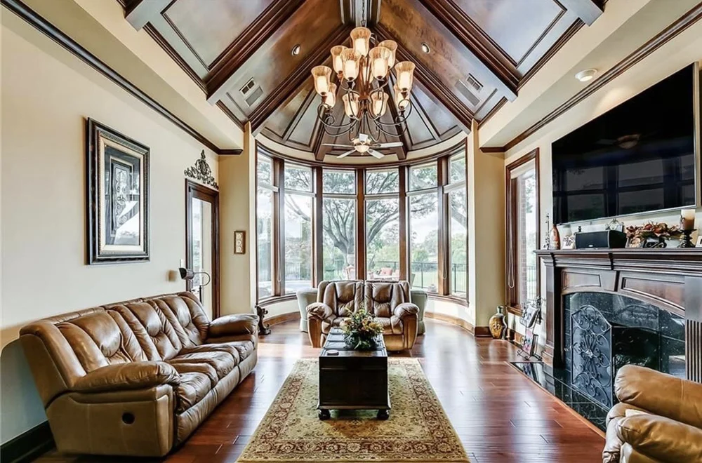 A spacious living room with large bay windows, a wooden vaulted ceiling, a chandelier, a fireplace, leather sofas, a flat-screen TV, and hardwood floors.