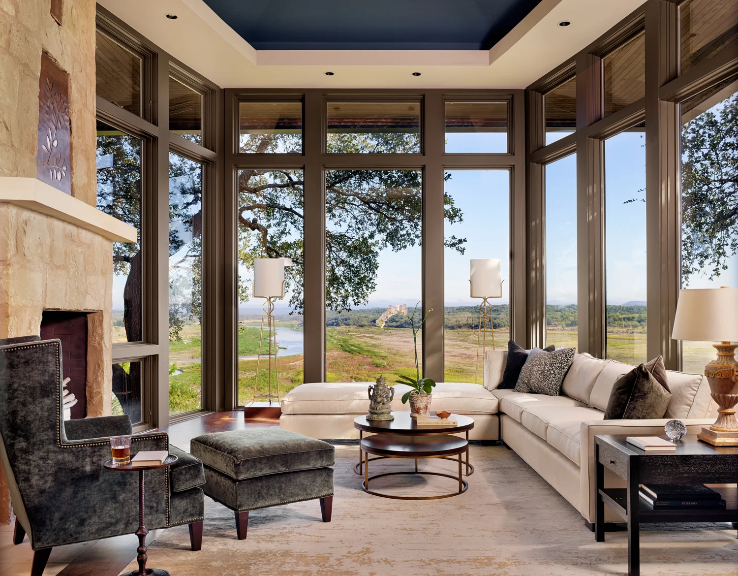 Living room with large windows overlooking a scenic landscape, featuring a white sectional sofa with dark and textured pillows, a dark armchair with an ottoman, a round coffee table with decorative items, and a side table with a lamp, against a stone