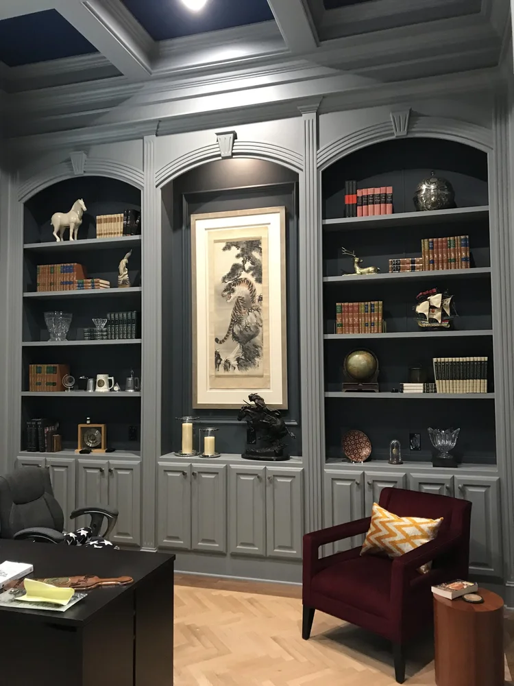 Interior of a sophisticated office or study room with built-in gray bookshelves filled with books, decorations, and art pieces, a gray chair, a burgundy armchair with a patterned pillow, and a wooden side table.