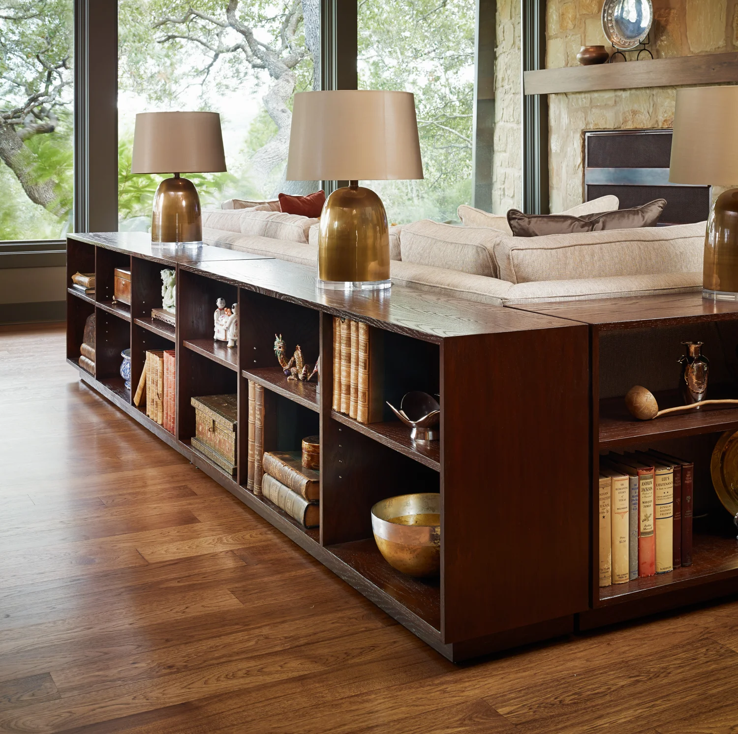 Living room with a wooden bookshelf topped by three beige lamps, beige sofa, stone wall, large windows showing trees outside, and hardwood floor.