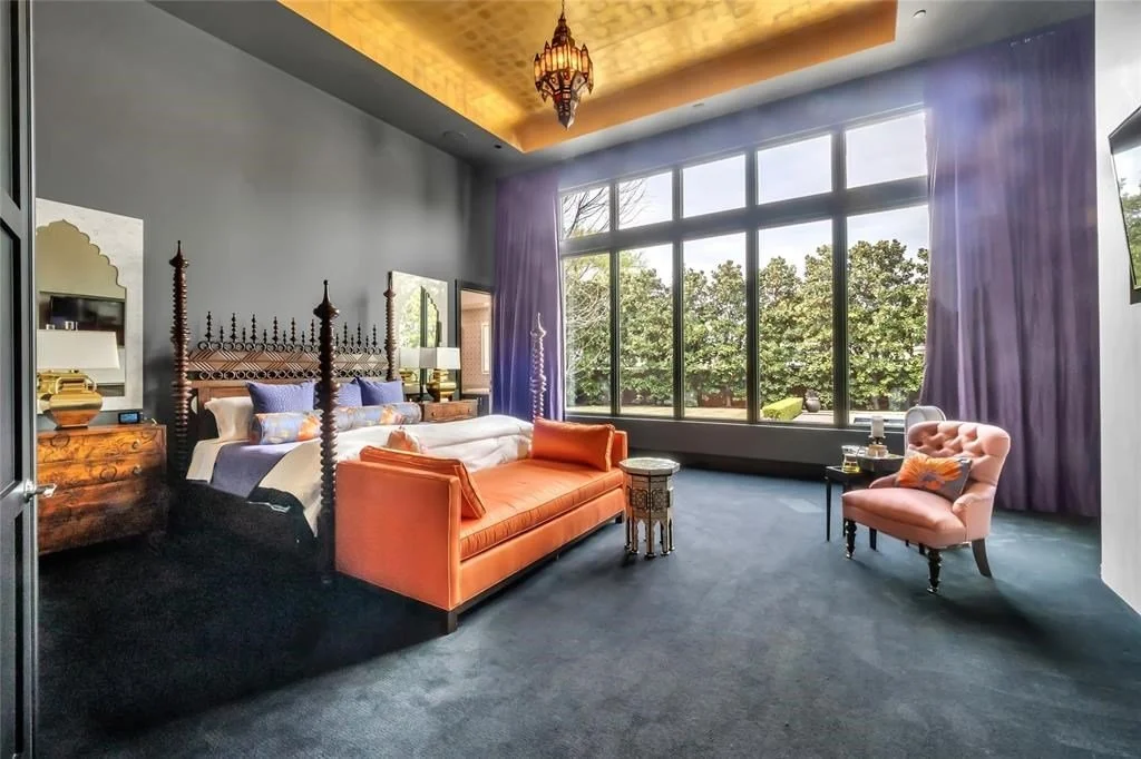 Elegant bedroom featuring a large bed with wooden posts, orange and purple accents, a big window with purple curtains showing greenery outside, a pink armchair, a small side table, and a decorative chandelier.