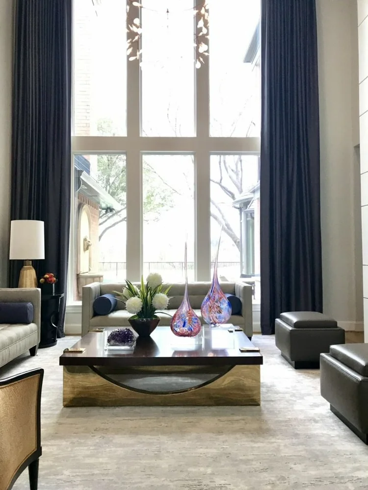 A living room with large window, navy curtains, sofa, armchair, coffee table with floral arrangement, and decorative glass vases.