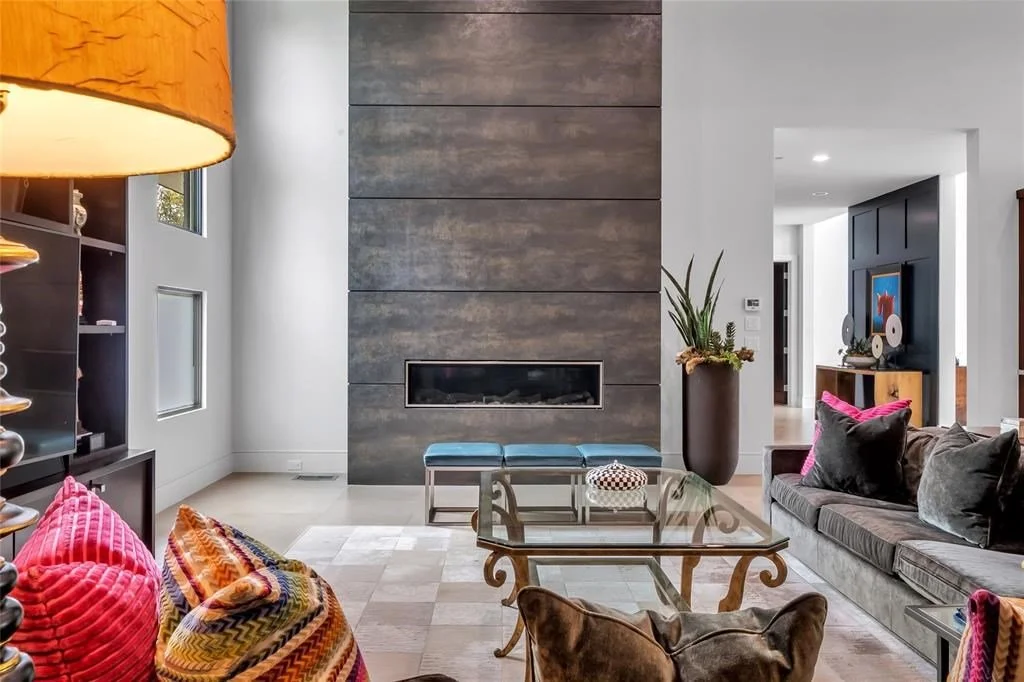 Living room with modern grey fireplace wall, glass coffee table, velvet sofa with colorful pillows, and large potted plant near the corner.