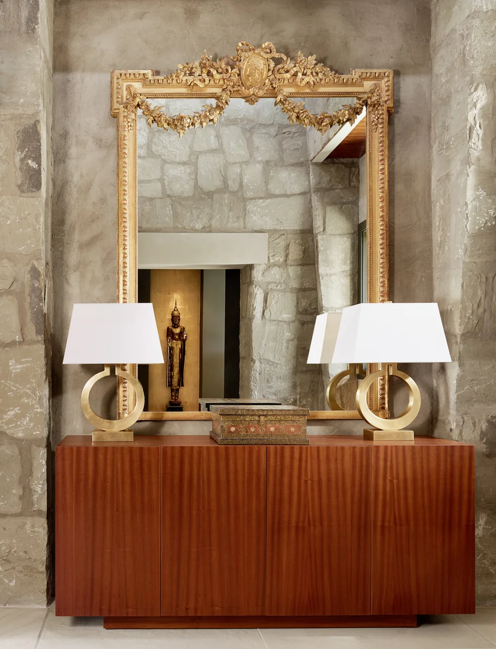 A wooden sideboard with two gold table lamps featuring circular bases, a mirror with a gold ornate frame and decorative garland, a small decorative box, stone wall background, and a reflection showing a Buddha sculpture in the mirror.