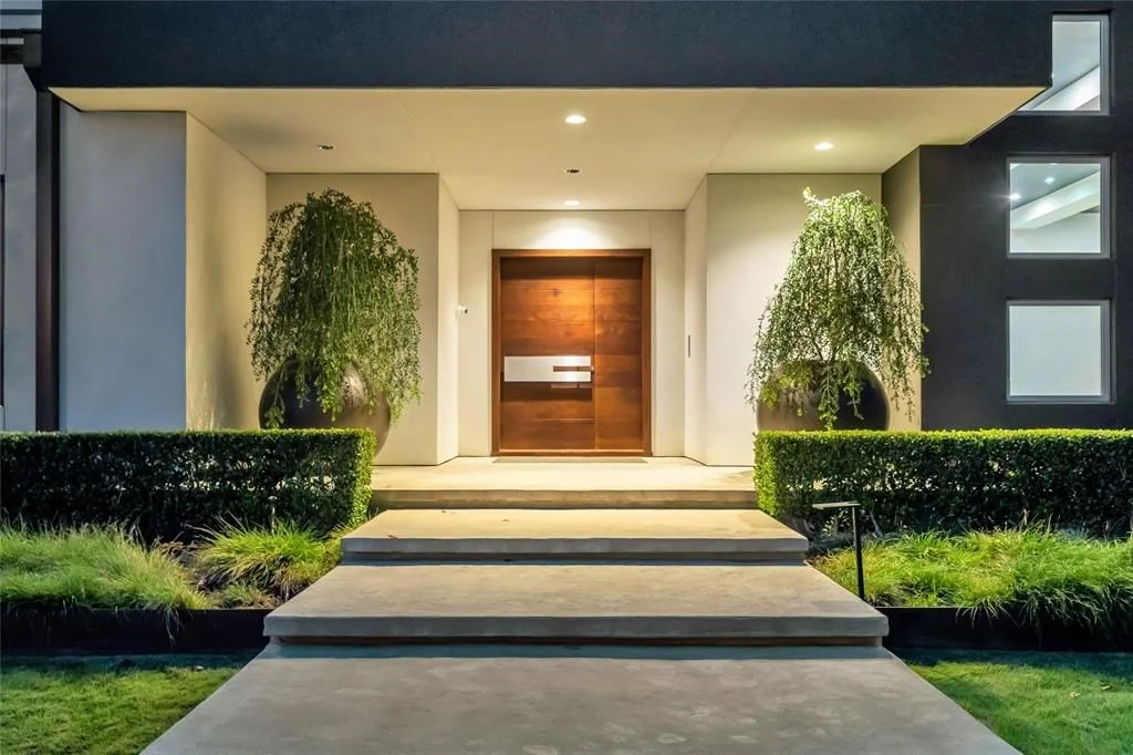 Modern house entrance with wooden door, manicured bushes, potted plants, and outdoor lighting