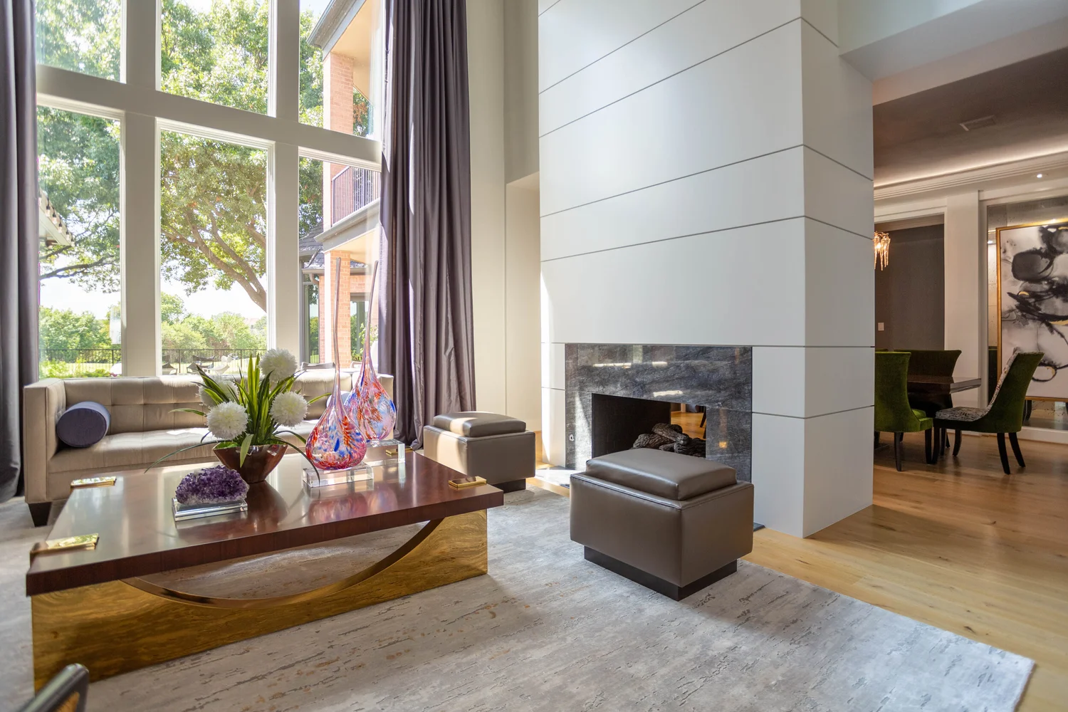 Living room with large windows, a beige sofa, a wooden coffee table with colorful glass vases and flowers, a modern fireplace, and a dining area with green upholstered chairs, all illuminated by natural light.