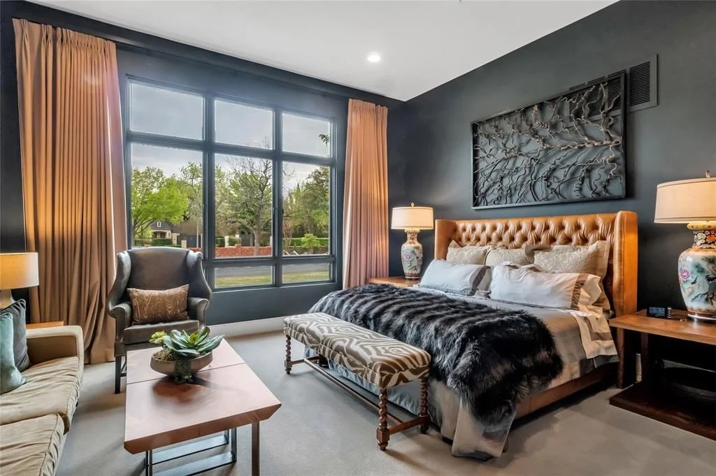 A cozy bedroom with a large window, a tufted leather headboard, and elegant decor including lamps, a bench, and artwork on the dark-colored wall.