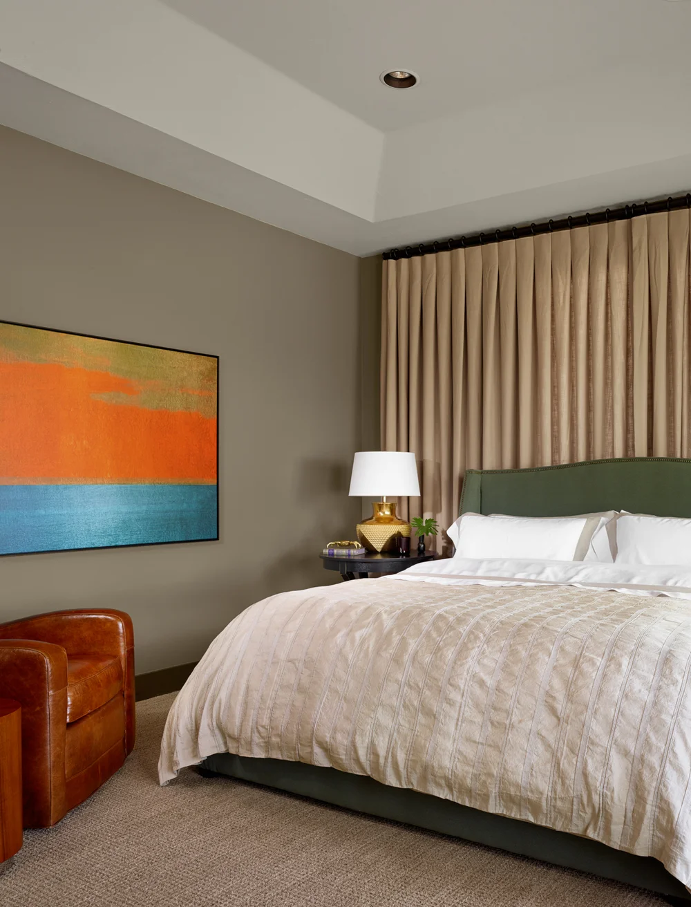 Hotel bedroom with a bed, green headboard, beige bedding, white pillows, a nightstand with a white lamp, a leather armchair, and a colorful abstract painting on the wall.