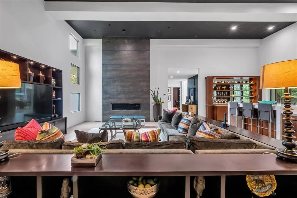 Modern living room with gray sofa, colorful pillows, black entertainment unit with TV, wooden bar with chairs, decorative vases, and a coffee table, with a fireplace on a gray accent wall.
