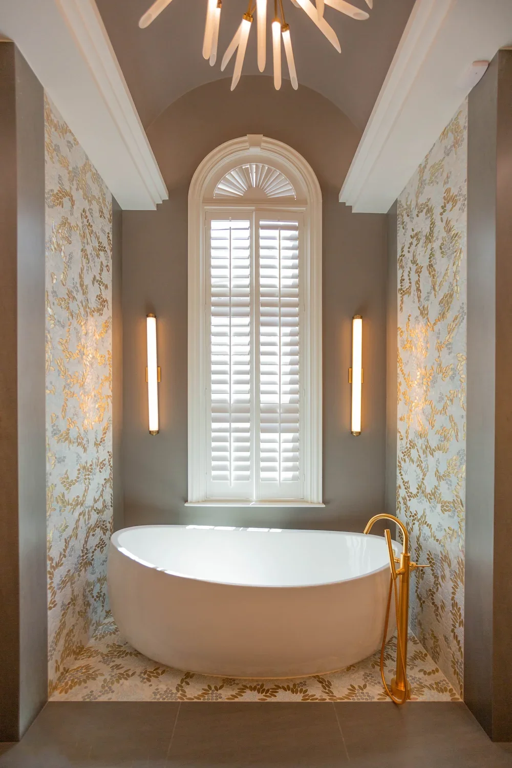 A luxurious bathroom with a white bathtub, gold fixtures, and a large window with shutters, floral wallpaper, and modern wall lamps.
