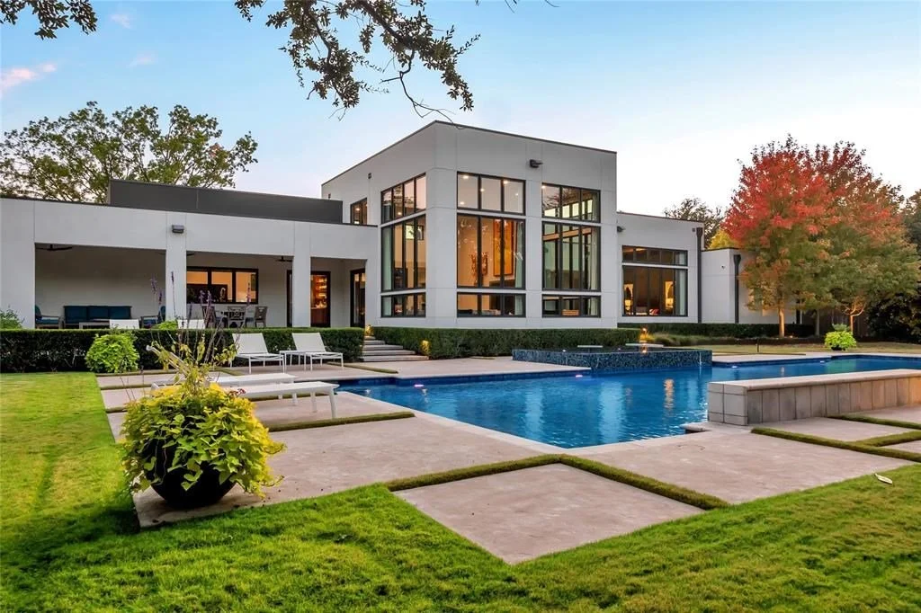 Luxury modern house with large glass windows, a rectangular swimming pool, and well-maintained lawn and landscaping, including potted plants and trees with autumn foliage.