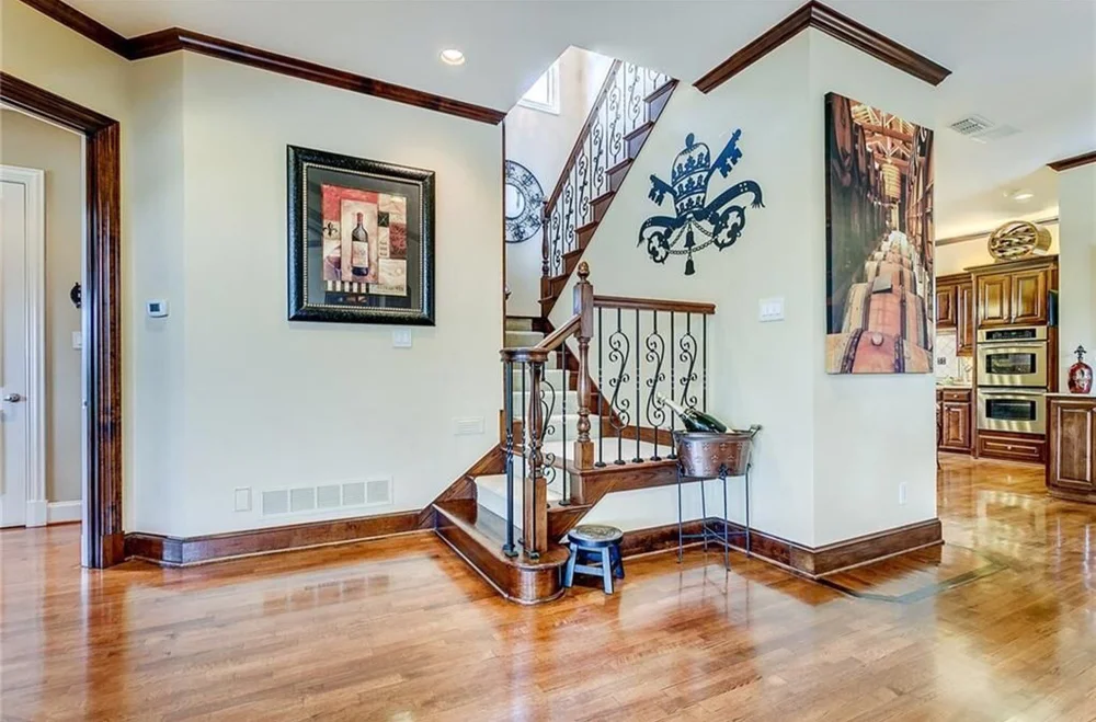 Interior view of a home entrance with wooden floors, staircase with black wrought iron railing, and wall decor including a framed picture and a wall decal of a crown and sword. The kitchen is partially visible in the background with wooden cabinetry.