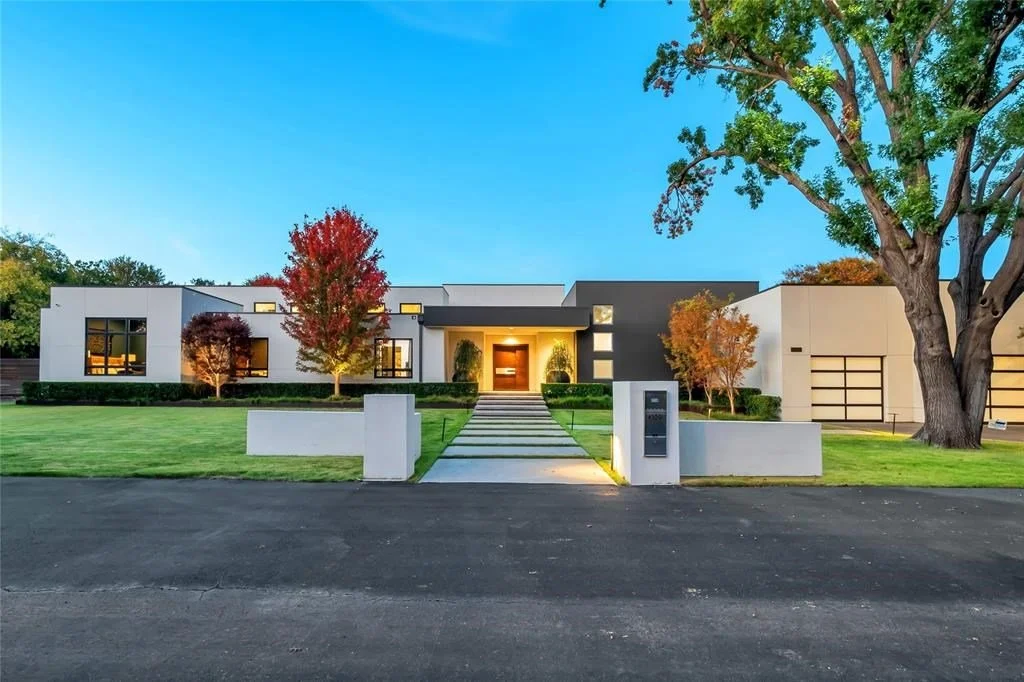 Modern single-story house with a white and gray exterior, front yard with green lawn, colorful trees, and a driveway with stepping stones leading to the front door during sunset.