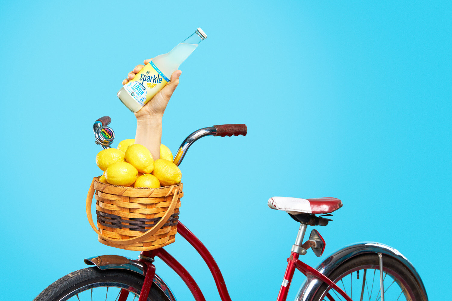 Quirky product photography: hand holding a bottle of lemon Sparkle by WiscoPop! emerges from a kids bike basket full of lemons.
