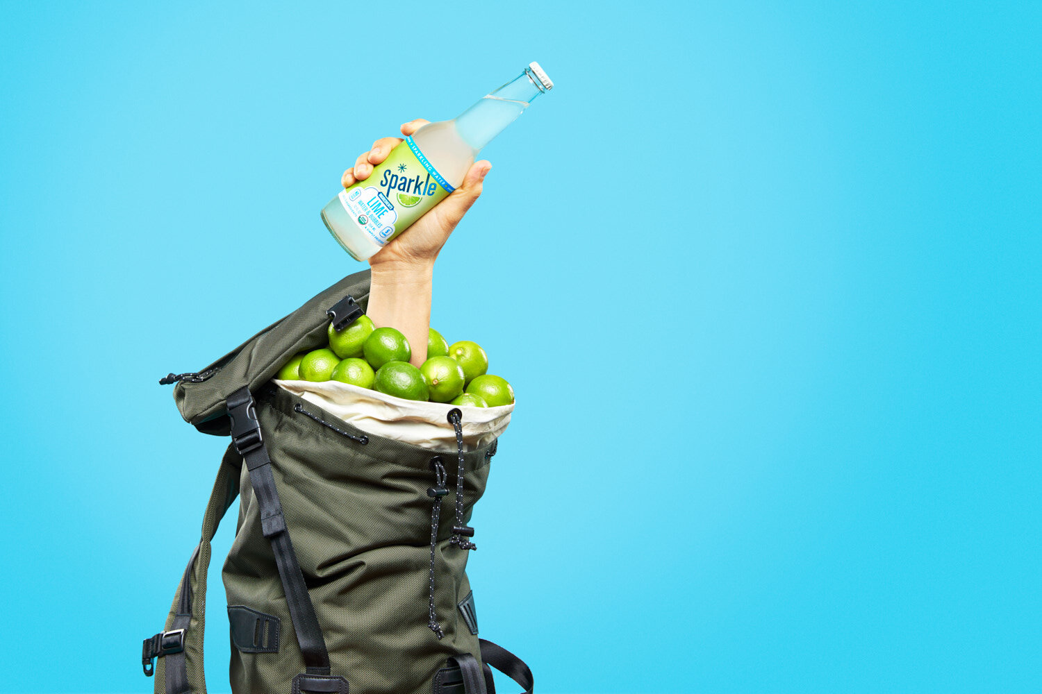 Quirky product photography:  hand holding a bottle of lime Sparkle by WiscoPop! emerges from a hiking pack full of limes. 