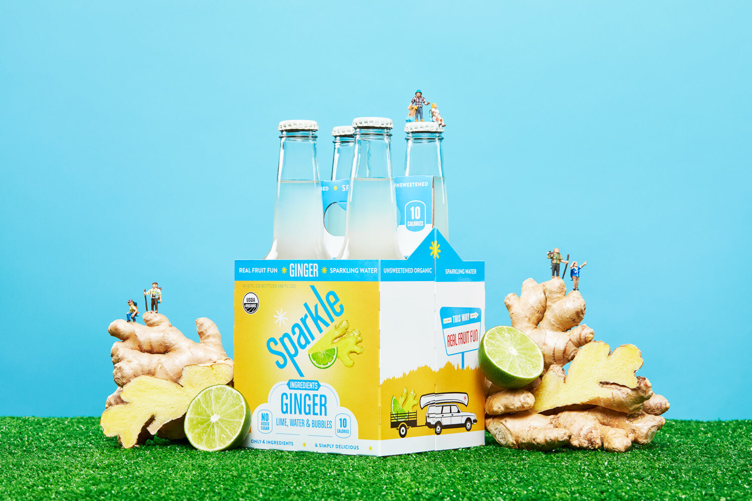 Creative product photography: four pack of ginger Sparkle by WiscoPop on grass with ginger root and limes.  Tiny figures of people hike the produce. 