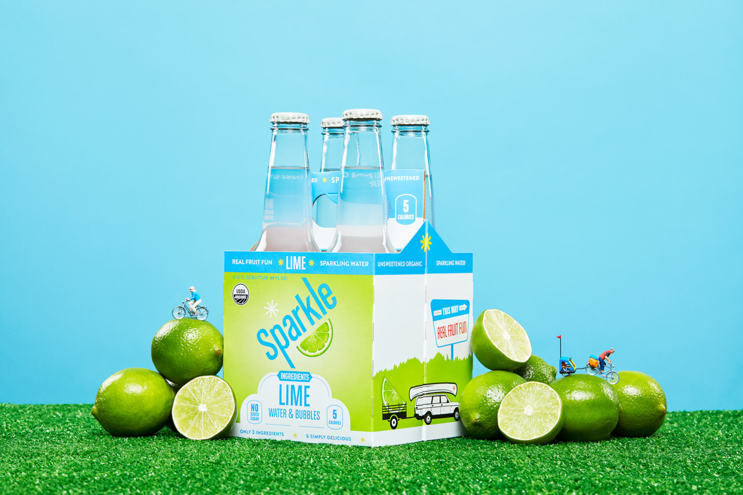 Creative product photography: Four pack of lime Sparkle by WiscoPop on grass with limes.  Tiny figures of people bike across the produce. 