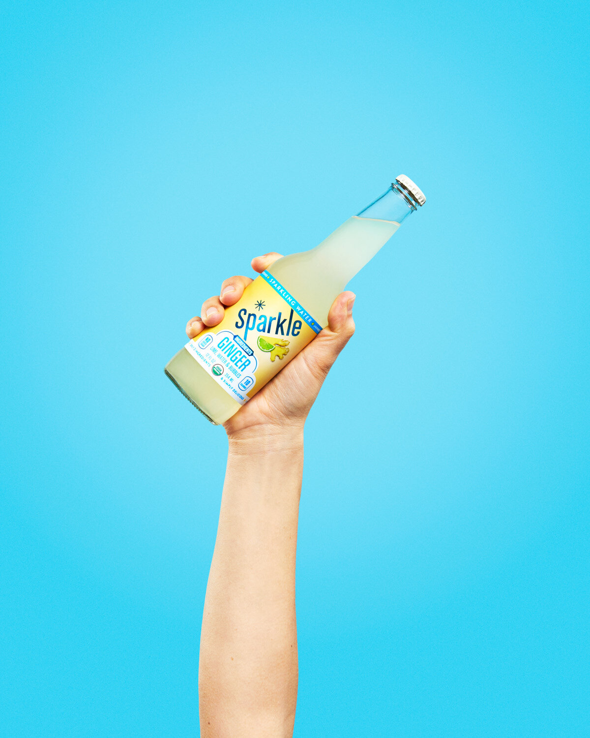 Colorful beverage photography: hand holds a bottle of ginger Sparkle by WiscoPop! on a blue background. 