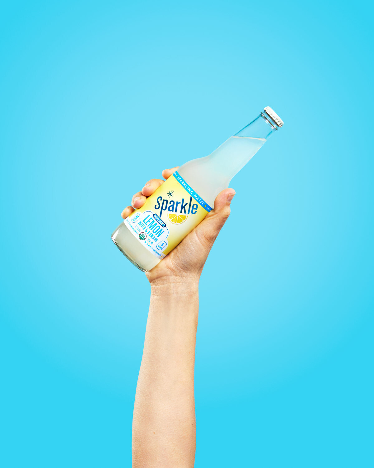 Colorful beverage photography: hand holds a bottle of lemon Sparkle by WiscoPop! on a blue background. 