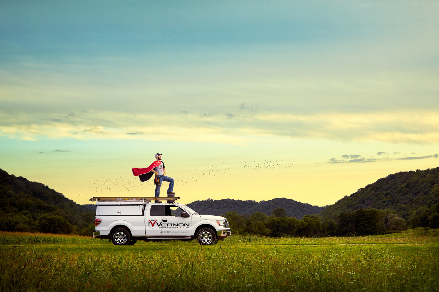 employee poses in cape as superhero on top of a truck in rolling hills of WI. for a creative advertising campaign by commercial photographer Hanna Agar