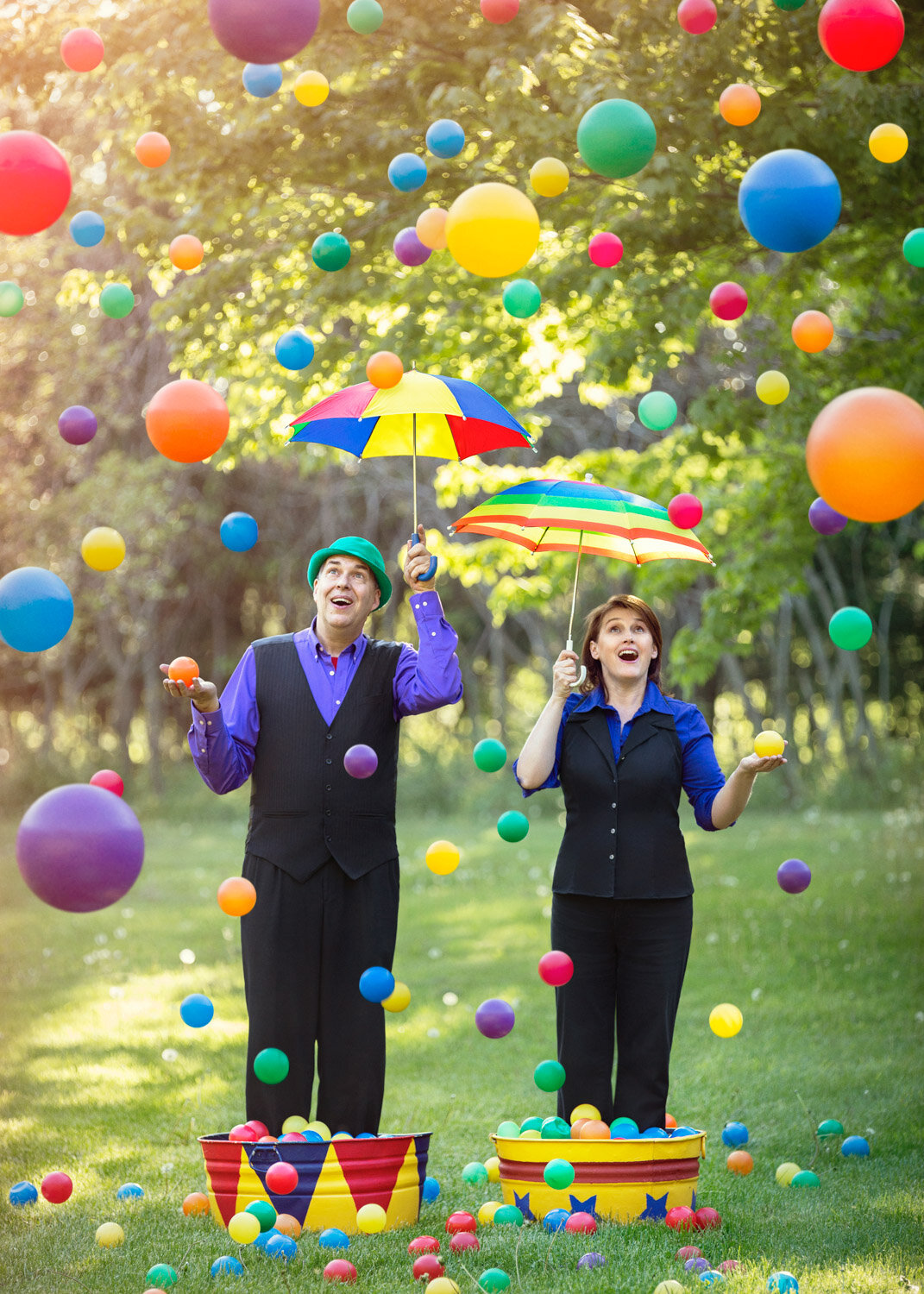 whimsical promotional photo of juggler duo standing in a kiddie pool with umbrellas and colored balls raining down by conceptual portrait photographer Hanna Agar